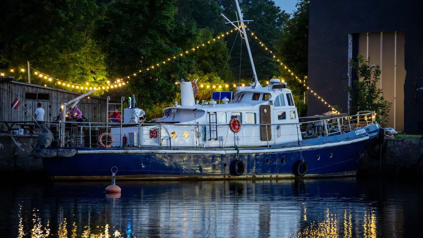 M/V LOTOSS lying alongside the quay at dusk, festoon lights strung from her mast and her reflection in the water.